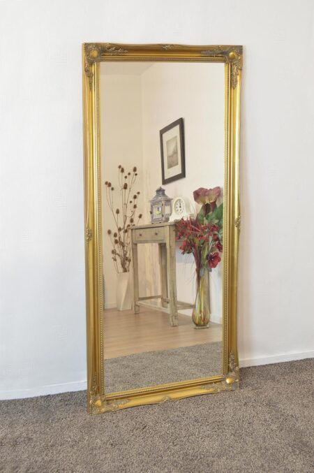 Full length mirror gold trim ornate frame leaning against a white wall in a carpeted room