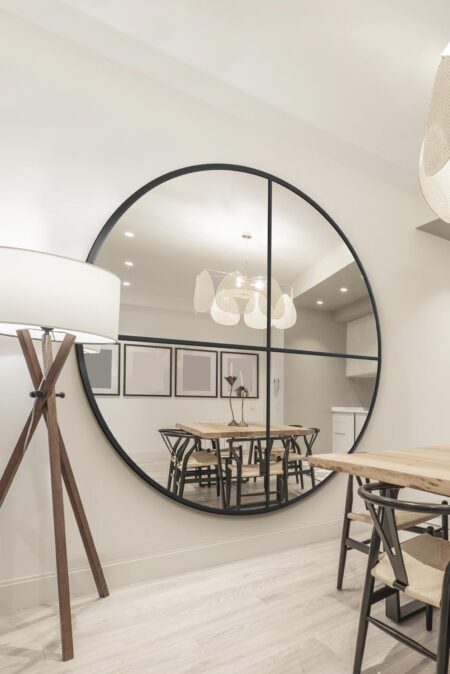 Large circular black-framed window-style mirror leaning against a wall in a bright dining room