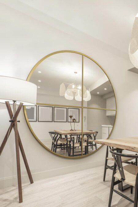 Oversized round gold crossbar mirror leaning against a white wall in a bright dining room