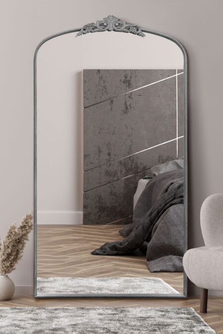 Full length silver wall mirror with ornate arch crest leaning against a bedroom wall