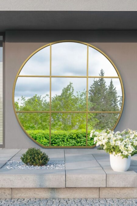 Extra large round gold metal mirror with nine-pane window grid mounted on an outdoor garden wall