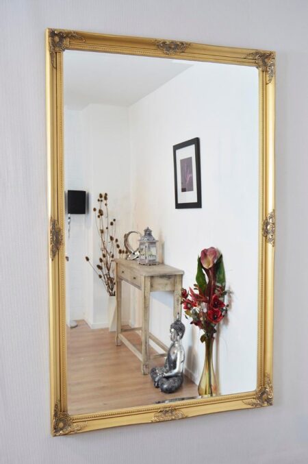 Gold ornate framed mirror hung on a wall reflecting a rustic console table and red flowers